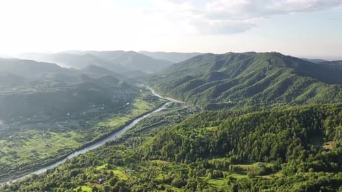 Flight over the summer mountains with mountain river serpentine, green meadows, village and forest.