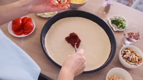 Woman Spreading Tomato Paste on Pizza Dough with Ingredients on Table Top View