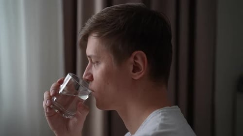Side View of Ill Young Man Taking Pills Drinking Fresh Water From Transparent Glass Standing By