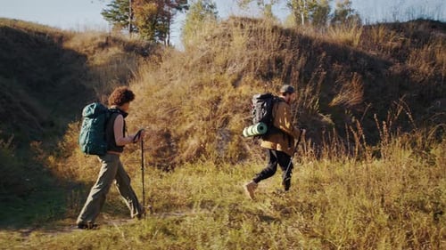 Young Man and Woman with Backpacks and Poles Walk Past Hills