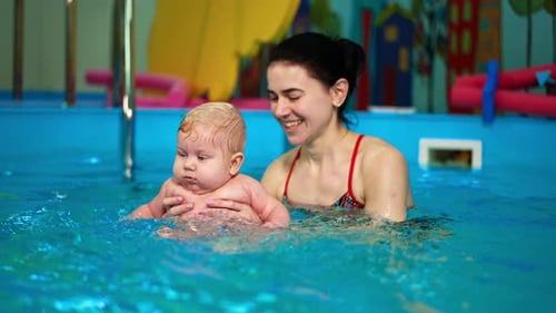 Adorable chubby baby is in the swimming-pool. Brunette woman teaching the infant to swim.