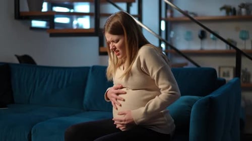 Junge Frau, die auf dem Sofa sitzt und starke Schmerzen im Bauch der Schwangerschaft hat, Porträt einer traurigen Kaukasierin