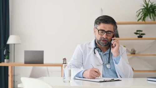Talking by phone and writing data. Mature male doctor in coat is in the clinic room