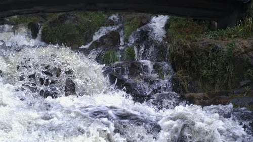 Low angle shot of a mountain stream cascading over rocks and vegetation under a bridge in Ultra