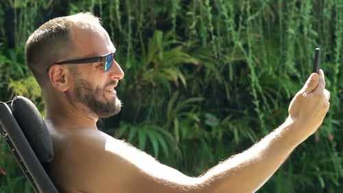 Happy man chatting on smartphone and drinking whiskey by luxury swimming pool