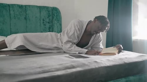 Man in Bathrobe Reading Book While Laying on Bed with Soft Light in Cozy Room