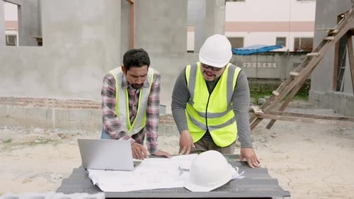 On-site serious standing construction workers actively discuss house plan. Asian engineers in yellow