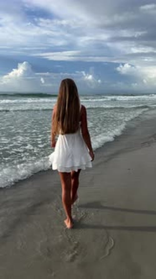 A Woman in a White Dress Walks Barefoot on a Serene Beach