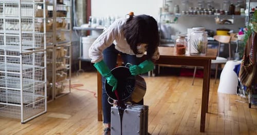 Female Craftsman Pouring Black Liquid From Pot Into A Mold For Soap Making In Interior Soap Works...