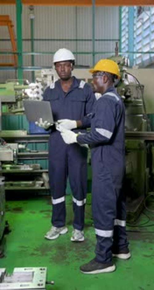 Two adult male technicians in hard hats review laptop data beside production equipment,