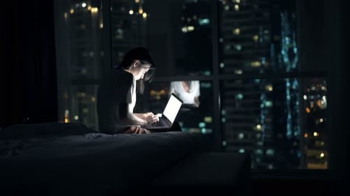 Woman Browsing Web On Laptop Sitting On Bed At Home At Night