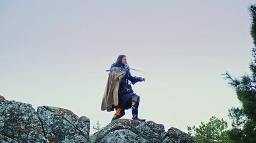 Un hombre disfrazado de caballero medieval con una espada está de pie sobre una piedra bajo los brillantes rayos del atardecer