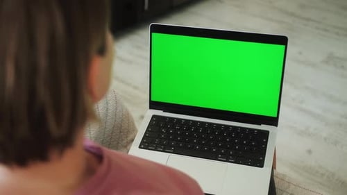 Woman Sitting on Sofa in Living Room and Looking at Laptop with Blank Green Screen