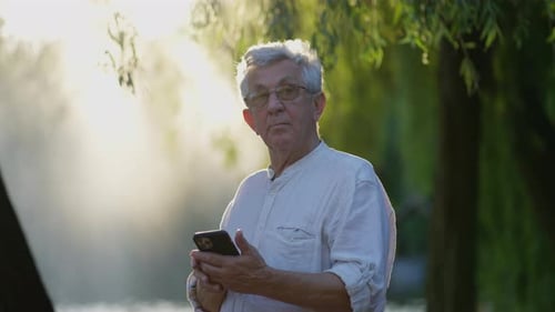 Senior Man Using Smartphone Outdoors Elderly Man with Glasses Focused on Mobile Phone in Sunlit Park