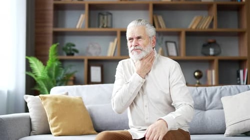 A sick elderly senior gray haired man suffering from a sore throat sitting on sofa in living room