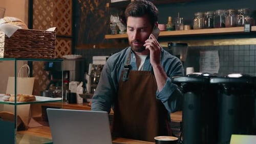 Man in Cafe Using Laptop and Talking on Phone