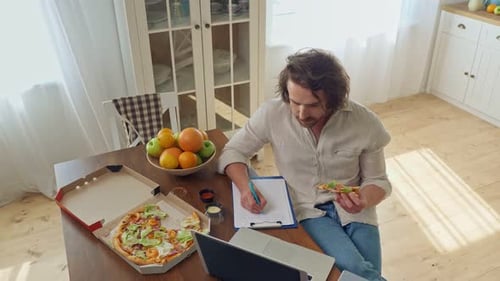Attractive Student Studying Remotely With A Laptop From Home Man Sitting at the Table Eat a Pizza