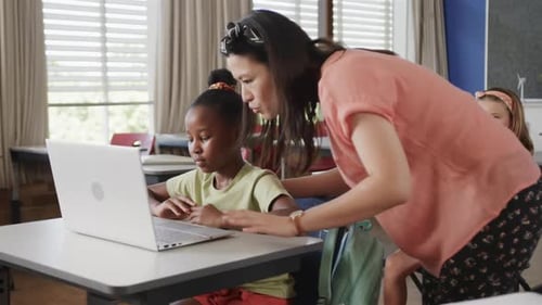 Happy diverse female teacher teaching schoolgirls using laptop in classroom at elementary school