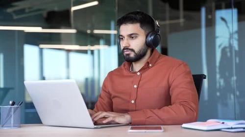 A focused IT specialist in wireless headphones works on a laptop sitting at a workplace in a busines