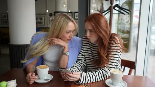 Portrait of Two Cheerful Girlfriends Women Sitting in Cafe Looking at Old Photos on Smartphone and