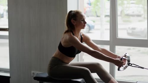A Girl Athlete in the Gym Near the Window Does Exercises Sitting on a Machine A Young Sportswoman