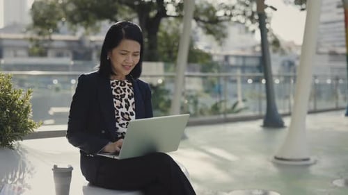 Asian Senior Office Woman Remotely Working on Laptop Chatting with Colleagues While Sitting Outdoors