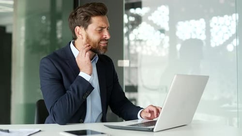 Upset businessman suffering from toothache working on laptop sitting at workplace at desk in modern