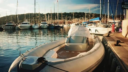 An inflatable motorboat tied to a dock in a busy marina, with sailboats and yachts reflected in calm