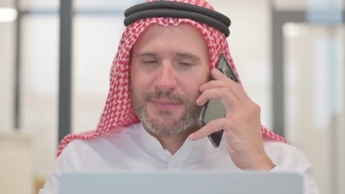 Man Using Laptop Answers Phone in Bright Office