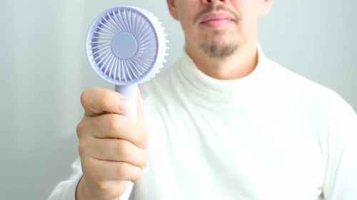 Young man holding mini fan on a gray background close-up, front view.