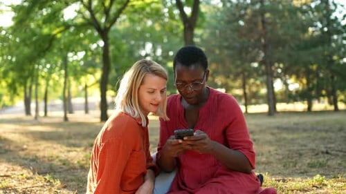Two diverse female friends sitting in a park using a smartphone and smiling together in sunlight