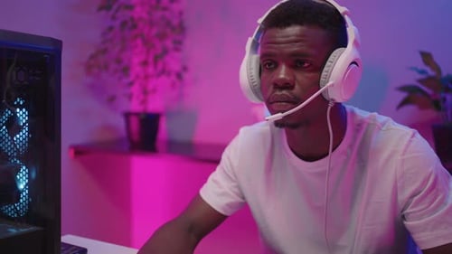 Porträt eines jungen schwarzen männlichen Spielers mit Headset, der alleine im Raum mit neonrosafarbenem Licht sitzt