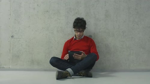 Young man students is sitting on the floor in a college hallway and using a tablet computer.