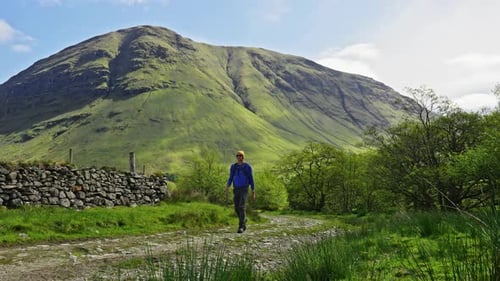 Hiker walking by on dirt path near stone fence and forest with grassy highland mountains in distance