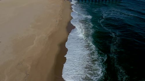 aerial flight along a beach with rippling waves tilts up to reveal a long pier dock