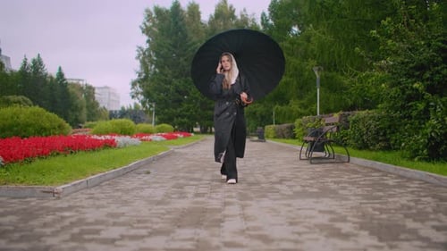 Maiden with Umbrella in Black Coat Walks on Wet Stone Path in Park