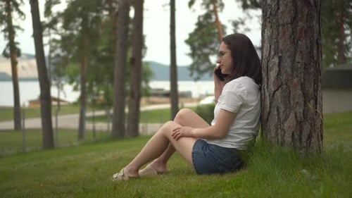 The Girl is Sitting on the Grass and Talking on the Phone A Young Woman in Glasses Sits Under a Tree