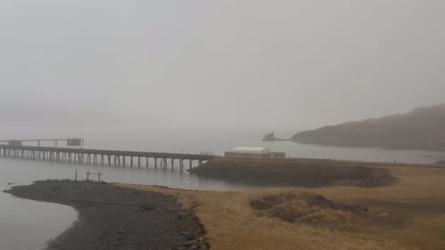Paisagem costeira nebulosa em Hvalfjörður, Islândia, com um longo cais de madeira que se estende sobre águas calmas,
