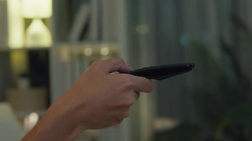 Nahaufnahme junger asiatischer Mann mit lässigem Stoff sitzt auf dem Sofa, hält die Fernbedienung gedrückt, schaut auf den Fernseher und wählt.