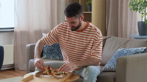Happy hispanic man enjoys tasty takeaway mediterranean pizza at home on sofa