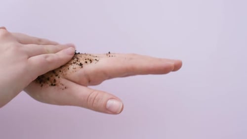 Young woman applies coffee scrub on her hands on pink background, side view.