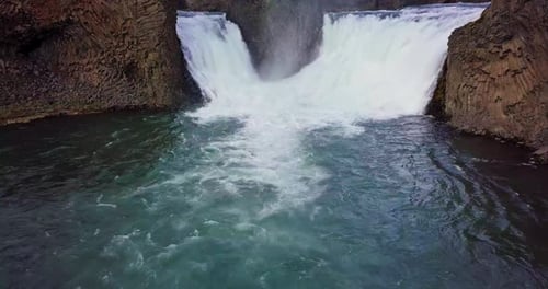 Static aerial of twin waterfalls flowing into a dark blue pond in Iceland.
