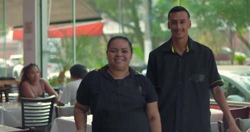 Latina waitress and waiter smiling at camera while standing together in dining room, light-filled