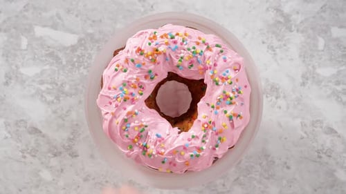 Flat Lay Sprinkling Rainbow Sprinkles Over Funfetti Bundt Cake Iced with Pink Buttercream Frosting
