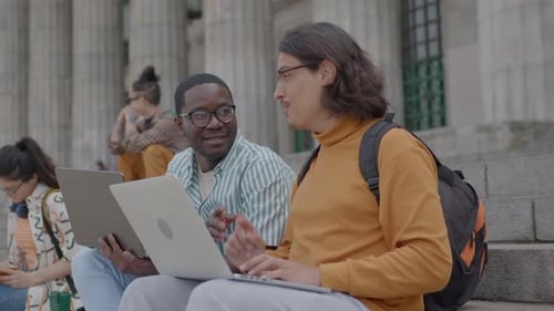 Two Groupmates Using Laptops and Talking on Stairs in front of University