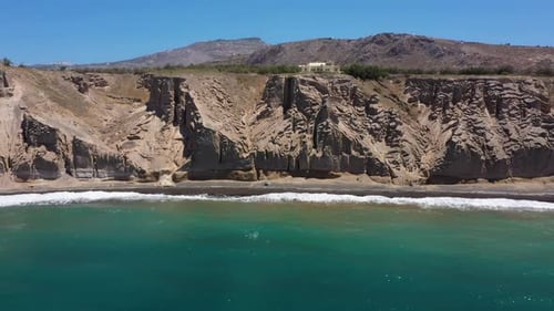 Aerial flying backwards of Greek Island, rocky cliff and blue water waves in the Mediterranean Sea o