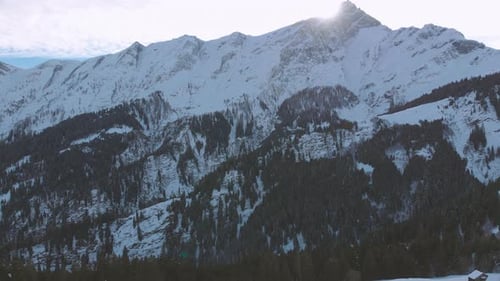 Ascending flying forward toward a huge mountain, snow covered hills, forest at wintertime on a sunny