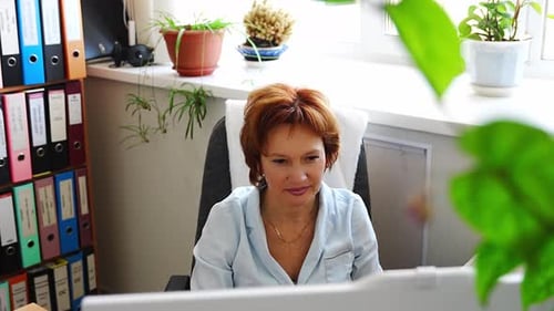 Woman Working at a Cluttered Office Desk with Folders Documents and Plants in the Background Concept