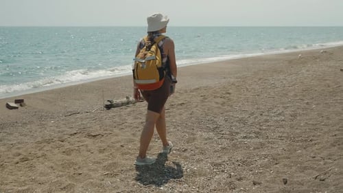 A Woman Walks Along a Quiet Beach With a Backpack, Enjoying the Gentle Sound of Waves and the Warm S