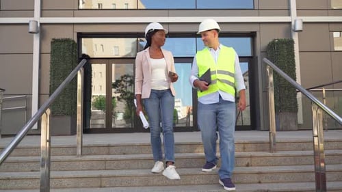 Two diverse construction engineers in hardhats discussing project while walking down stairs outdoors